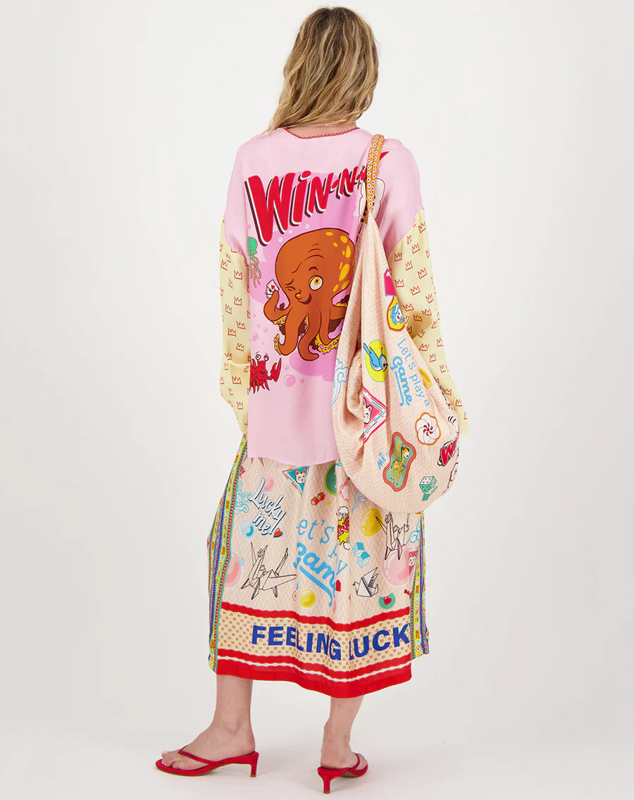 A woman faces away, showcasing a colorful, eclectic ensemble featuring patterns and text like "WINNER" and a cartoon octopus on her matching shirt. She wears the Vanessa Midi Skirt by Me369 with an elastic waist, carries a patterned bag, and completes her look with red sandals.