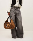 A person wearing BA&SH’s Jean Yusc—high-waisted, wide-leg gray jeans—pairs them with a white blouse and black vest, holding a brown suede shoulder bag. The image is cropped at the shoulders.