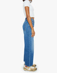 A woman with medium skin tone and curly hair stands sideways against a white background, wearing The Dodger Ankle high-waisted blue cotton blend loose straight leg pants by Mother, a white cropped tee, and worn beige Nike sneakers.