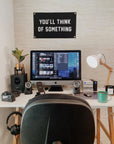 A tidy home office features an iMac, speakers, headphones, camera, plant, lamp, mug, and the Oxford Pennant "You'll Think of Something" Camp Flag 14x24—perfect for sparking inspiration and boosting creativity.