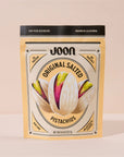 A beige resealable bag of Faire Joon Pistachios features an illustrated pistachio and highlights 6g protein, non-GMO, no artificial flavors, and no seed oils for a pure snacking experience.