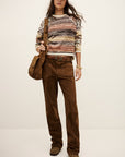 A woman wears the BA&SH Jumper Clodie— a wool sweater with brown-toned stripes—paired with brown suede pants, brown shoes, and a matching brown shoulder bag, standing against a plain light background.