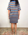 A woman with curly hair stands on a patterned rug, facing away from the camera, wearing the Xirena Justine Dress—a super-soft striped cotton jersey t-shirt dress in brown, blue, white, and yellow—and light-colored sandals.