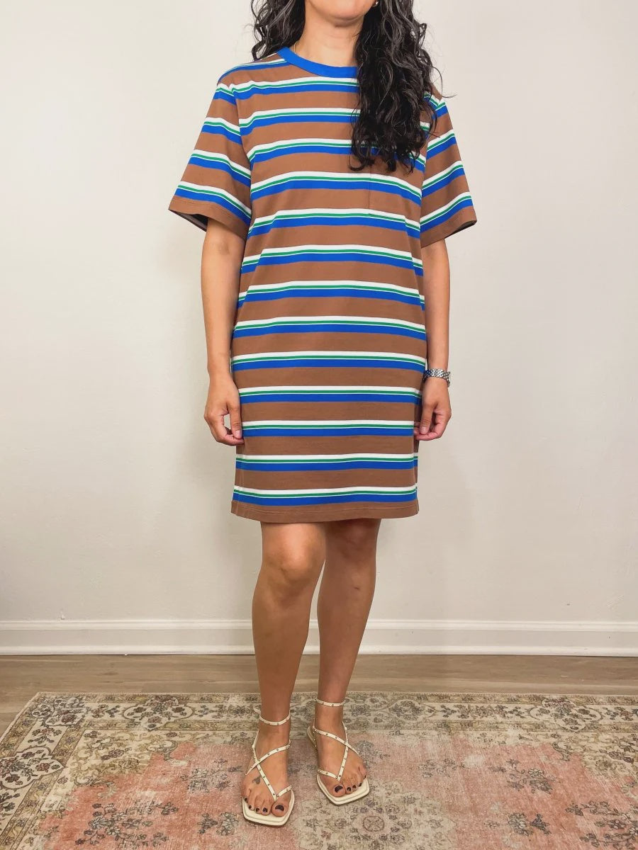 A woman stands indoors in Los Angeles wearing the Xirena Justine Dress, a brown, blue, and white striped cotton jersey t-shirt dress, paired with beige pearl-accented sandals. She stands on a rug against a plain wall.