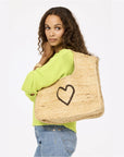 A woman with long curly hair in a light green sweater and blue jeans carries the Kerri Rosenthal Imperfect Heart Jute Tote, a large handmade bag with a black heart design, while standing against a plain light background.