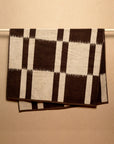 The Karin Bath Sheet by Autumn Sonata, with a large black and beige checkered pattern, is shown hanging neatly over a horizontal rod against a neutral beige wall.