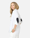 A woman with long blonde hair wears the Kerri Rosenthal Mia Shirt Classic White with black elbow patches and white pants. Smiling, she turns slightly to the side, her rolled-up sleeves accessorized with bracelets and rings.