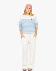 A woman faces forward, smiling slightly with her hands in her pockets. She wears a pink bandana, loose white pants, sandals, and the Kerri Rosenthal Artist Long Sleeve Stripe Tee—an oversized cotton shirt with blue stripes and a red heart on the chest.