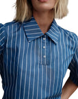 A person with blonde hair wears The Polo Shirt by Cissa, a blue collared top with white Keswick stripes and a partial button placket. The image is cropped to show only the head and upper torso.