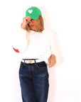 A woman in a white sweater and dark jeans adjusts her KR Imperfect Heart Hat from Kerri Rosenthal, featuring its signature exclusive color and white heart. Her "LOVE" belt accentuates her effortless chic pose against the plain white backdrop, embodying a timeless KR staple.