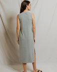 A woman with straight brown hair wears the Perfectwhitetee Lanell Cotton Tank Dress—a relaxed fit, sleeveless light gray dress with a side slit—paired with black sandals. She stands facing away, against draped, light-colored fabric.