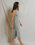 A woman with long brown hair wears the Perfectwhitetee Lanell Cotton Tank Dress, a relaxed fit gray midi tank style, along with black flip-flops as she walks in profile against a light fabric backdrop.
