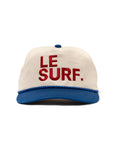 The Le Surf Vintage Lesure Icon Cap by Ping Pong Surf Club is a white cotton snapback with a blue flat brim and button, featuring "LE SURF." embroidered in bold red letters on the front.