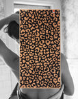 A person, back to the camera, stands in a bathroom wrapped in the Typical Wild Leopard Bath Towel. The bold leopard print towel from Typical drapes over their upper body, partially obscuring the view.