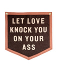 The Faire Let Love Knock You Camp Flag features a brown banner with a pink border, gold grommets, and bold cream letters reading "LET LOVE KNOCK YOU ON YOUR ASS"—relationship advice reminiscent of a Jason Isbell lyric.