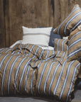 A person sits on a bed with brown and blue striped bedding, holding two matching pillows. Wooden paneling and the Libeco Home Salem Duvet Cover Stripe Twin create a cozy atmosphere, promising a comfortable sleep.