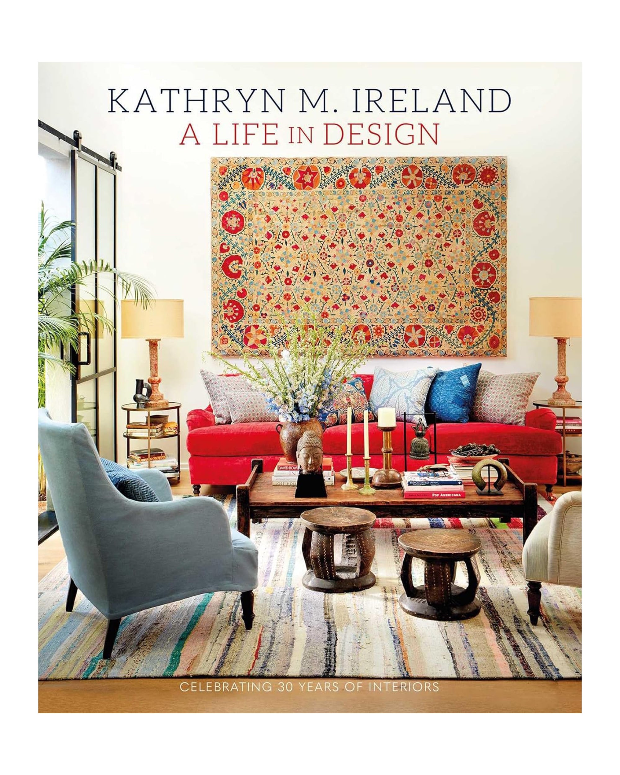 The cover of "Life In Design" by Simon & Schuster Inc. displays a vibrant living room—red sofa, patterned tapestry, blue armchair, eclectic cushions, lush plants, and wooden tables on a striped rug.