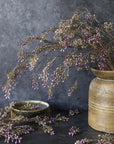The Berry Branch Lilac by Abigail Ahern sits in a rustic ceramic vase, its purple buds and artificial berries creating a striking scene. Some branches and berries are scattered nearby on the dark surface with a small bowl, set against a textured gray backdrop.