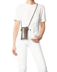 Wearing a plain white t-shirt and white jeans, a person stands with one hand in their pocket, modeling the Jerome Dreyfuss Louis Mobile Holder—a small metallic crossbody bag with an adjustable shoulder strap.