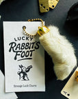 The Faire Lucky Rabbit's Foot keychain features a white rabbit’s foot with a gold cap, attached to a card that reads “Lucky Rabbit’s Foot, Strange Luck Charm,” with playful artwork and surrounded by cards and dice.