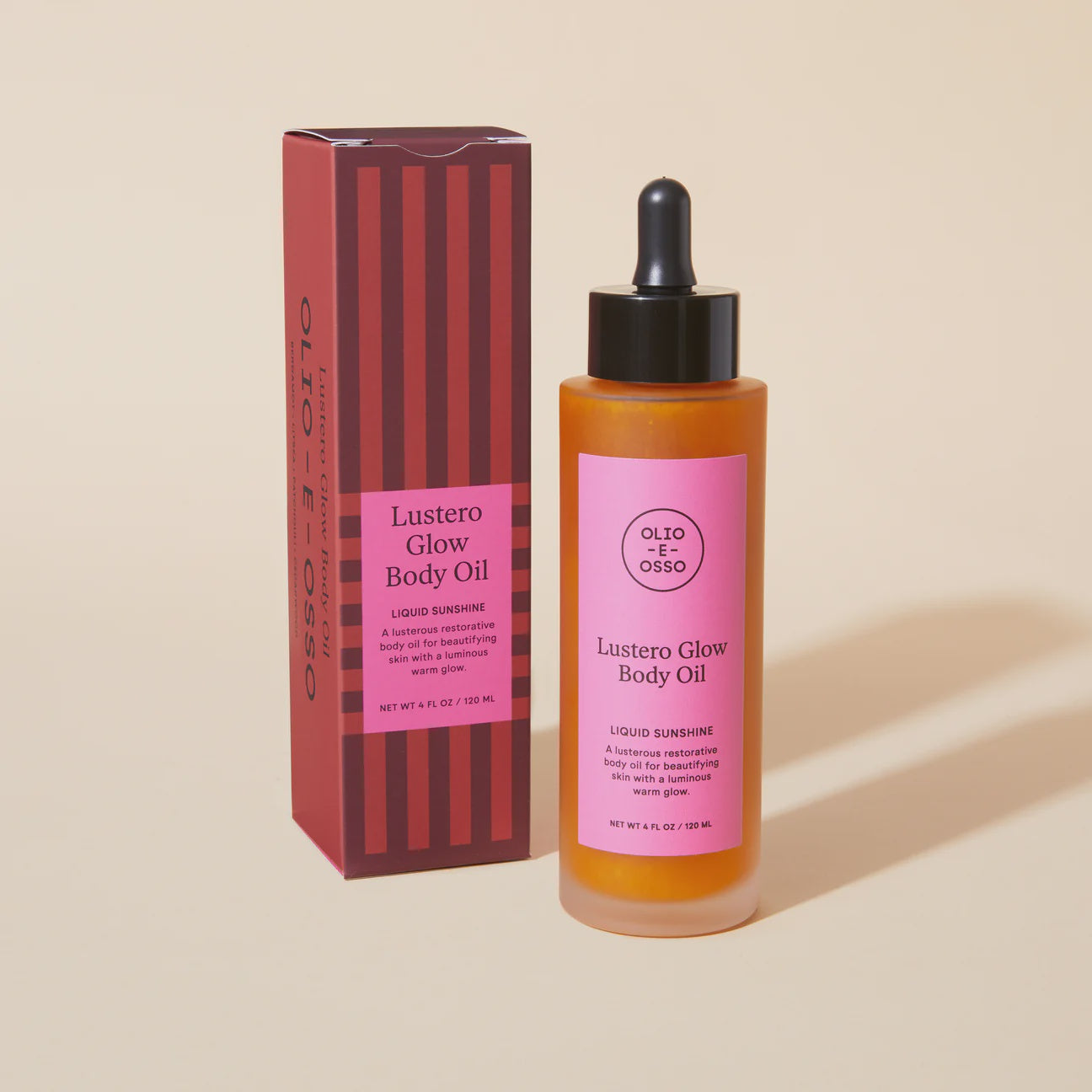 A bottle of Olio E Osso Lustero Glow Body Oil from Scottsdale, Arizona, next to its packaging. The bottle is amber with a dropper and the box is red with vertical pink stripes. Both items display the product name and details.