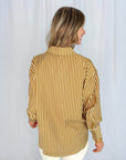 A person with blonde hair, seen from the back, wears the SUNCOO Lynsha Blouse—a loose-fitting, long sleeve collared top with vertical mustard yellow and white stripes—against a plain white background.