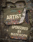 The limited-edition Artist Tote by Made Worn is crafted from cotton canvas with paint splatters, polka dot ribbons, confetti accents, and playful pins—one says "ARTIST," the other "SPONSORED BY MADEWORN." Each bag is truly unique.