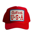 The Faire Made in the USA Patch Trucker Hat is a red cap with an adjustable snapback, blue trim, and a rectangular patch reading “Made in the U.S.A.” in red letters, accented by small red hearts between the letters.