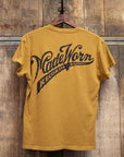The MadeWorn Records unisex vintage pocket tee by Made Worn features mustard yellow distressed fabric and "RECORDS HOLLYWOOD, CA." text on the back. It hangs on a wooden hanger against a rustic wall and is made in Los Angeles.