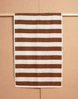 The Maria Hand Towel by Autumn Sonata, featuring a jacquard weave and brown and white horizontal stripes, is draped neatly over a wooden rod against a beige background.