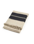 The Marshall Throw by Libeco Home is a folded beige wool blanket featuring a wide black stripe and flax fringe on one edge, displayed against a white background.
