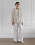 A person stands indoors against white paneled walls and a wooden floor, wearing LATIERRA's handcrafted Medusa Cardigan Coat—white and fluffy—paired with white wide-leg pants, a white top, and white shoes.