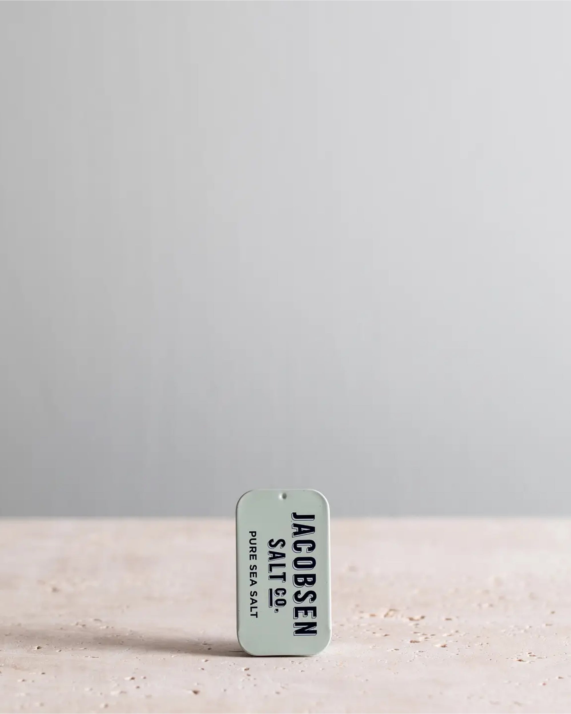 A small rectangular tin labeled "Faire Kosher Sea Salt slide Tin" stands upright on a light textured surface, offering convenient, travel-sized kosher sea salt against a plain, light gray background.