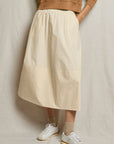A person wears the Perfectwhitetee Poplin A Line Skirt with pockets, paired with a tan sweatshirt, beige socks, and white sneakers. They stand against a light fabric backdrop with hands casually tucked in the skirt’s pockets.