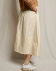 A person stands sideways in a tan sweatshirt, Perfectwhitetee Poplin A Line Skirt in cream with pockets, beige ankle socks, and white sneakers against a light fabric backdrop.