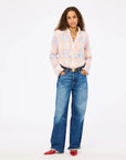A woman with long curly hair wears the Kerri Rosenthal Molly Shirt, styled with blue wide-leg jeans, red pointed-toe shoes, and layered necklaces. She stands with hands in her pockets against a plain white background.