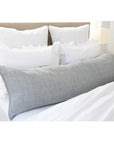 The POM POM Montauk Body Pillow in light gray heavy weight linen rests across a neatly made bed with white sheets and four ruffled white pillows set against a wooden headboard.