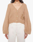 A woman wears Mother’s The Puffy Sleeve Varsity Cardi, a tan alpaca-blend knit with a chunky, cropped fit, large sleeves, three buttons, textured pattern, and an embroidered detail on the left side, paired with high-waisted white pants.