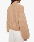 A woman with dark hair models Mother’s The Puffy Sleeve Varsity Cardi—a textured, loose tan sweater with balloon sleeves—paired with high-waisted white pants, shown from the back against a plain white background.