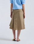A person stands facing away, wearing a light blue short-sleeve shirt, tan sandals, and the Rachel Comey Muci Skirt—a high waisted khaki skirt with ruched sides—against a plain light background.