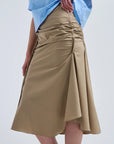 A person wearing a light blue shirt and the Rachel Comey Muci Skirt—a high-waisted, beige ruched knee-length skirt with a front slit—stands against a plain light background.