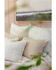 A cozy bed is adorned with a variety of white and beige textured pillows, including the luxurious Murphy Big Pillow 28x36 from POM POM, some of which feature a plush bouclé texture. A soft pink wall forms the background, showcasing a framed abstract painting depicting a serene landscape. The foreground includes blurred green plant leaves.