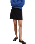 A person wearing a textured blue sweater, the Alex Mill Naomi Mini Skirt in black with side zip closure, and black loafers stands with arms crossed against a plain white background.