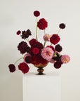The Naomi Compote by Accent Decor displays dark red, bright red, and light pink dahlias with long stems in a brown vase, set on a white pedestal against a plain white background.