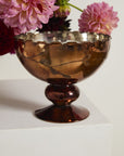 The Naomi Compote by Accent Decor features bronze-colored mercury glass with a scalloped rim, set on a white pedestal and filled with pink and purple dahlias.