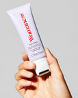 A hand holds a white tube labeled “Faire Let’s Neck – Neck Serum w/ Cooling Applicator” against a plain gray background, featuring red and black text and a cooling roller tip for neck and décolleté care.