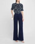 Woman wearing the Apiece Apart Los Altos Top in navy with an abstract print, tucked into high-waisted wide-leg navy pants, paired with a small white handbag and white heeled sandals.