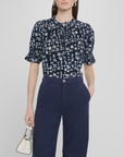 A woman stands against a plain light background wearing the Apiece Apart Los Altos Top, a navy blue short-sleeve blouse with a crew neckline and floral print, tucked into high-waisted navy pants and holding a small white handbag.