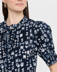 A woman wears the Apiece Apart Los Altos Top in navy blue with an abstract print, ruffle collar, and puffed short sleeves. She accessorizes with gold rectangular earrings. Only her head and upper torso are shown.