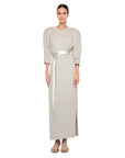A woman stands barefoot against a white background wearing the Norma Kamali Bomber Long Dress—a vintage-inspired, light gray dress with three-quarter sleeves, subtle puffed shoulders, and a shiny silver belt at the waist.
